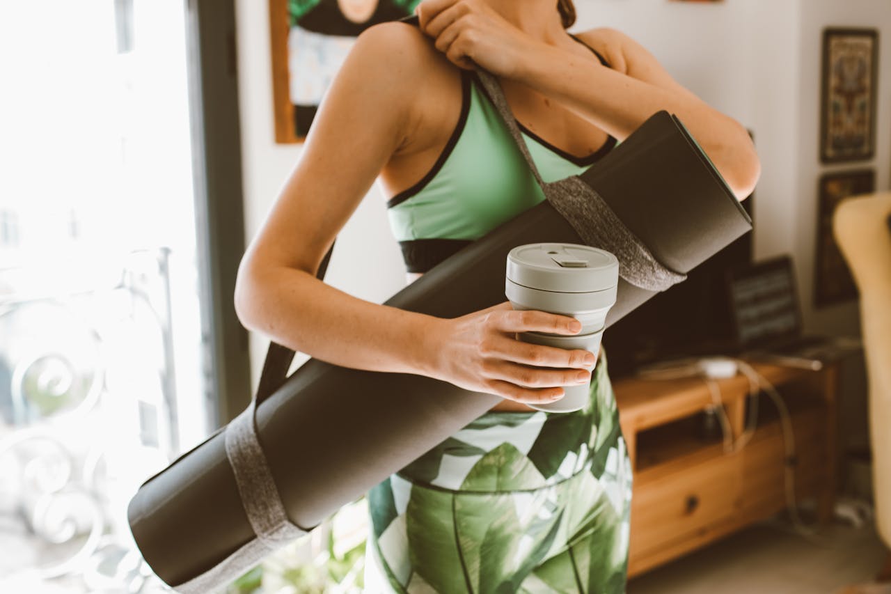 Young woman in sportswear holding yoga mat and cup indoors, ready for workout.